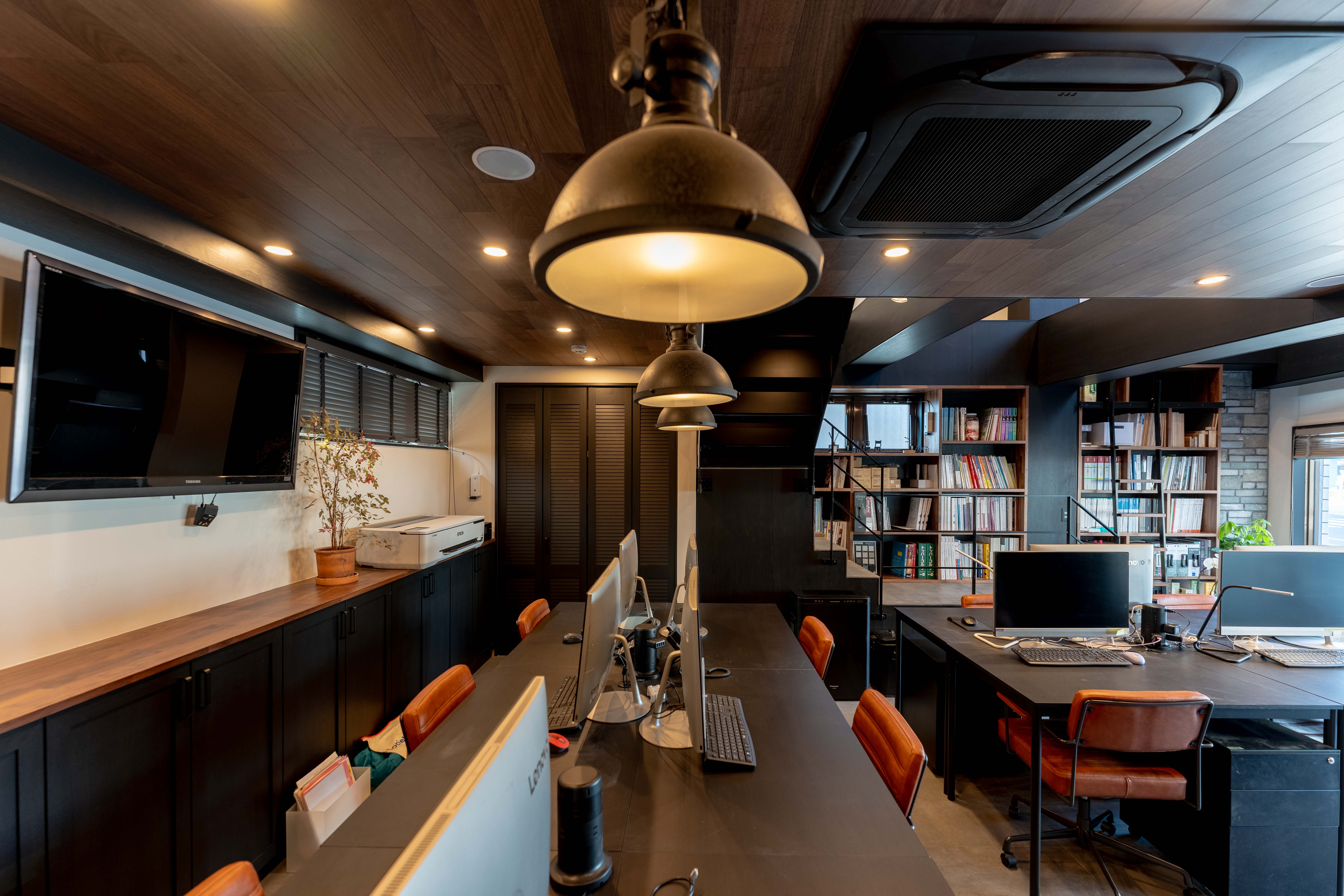 ラトリエの事務所の写真。木張りの天井から金属製のペンダントライトが吊るされたオフィスで、中央には黒い長机とデスクトップパソコン、オレンジブラウンのレザーチェアが並び、奥には黒い階段と資料が並んだ大きな本棚が見える。