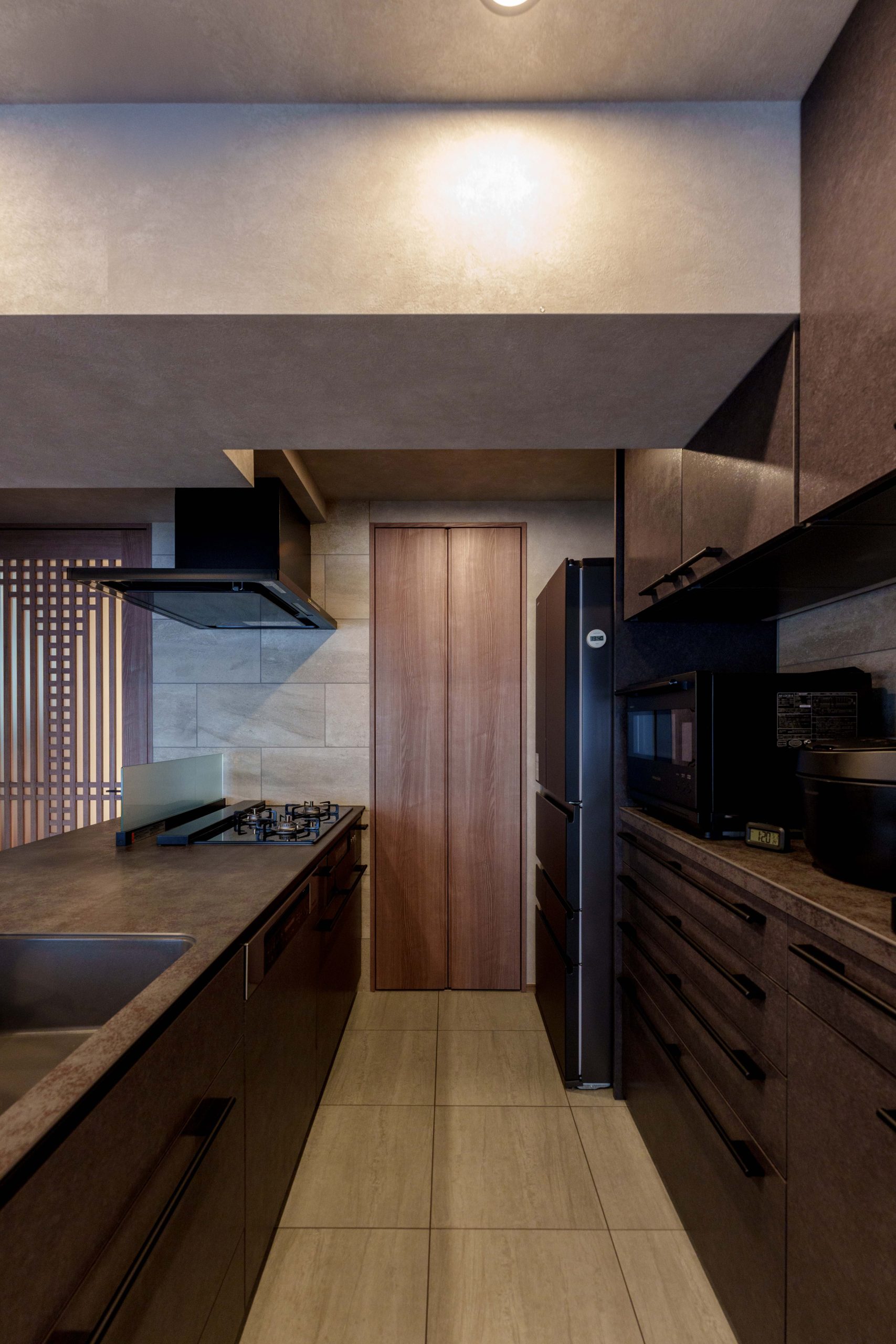 施工写真。重厚感のあるダークグレーのワークトップとキャビネットを採用したキッチン空間。左手にはガスコンロとシンク、右手には家電が並ぶカップボードと冷蔵庫が配置されている。正面の壁はグレーのタイル張りで、木目調のドアがアクセントになっている。照明を抑えた落ち着いたトーンが、都会的で洗練された雰囲気を演出している。