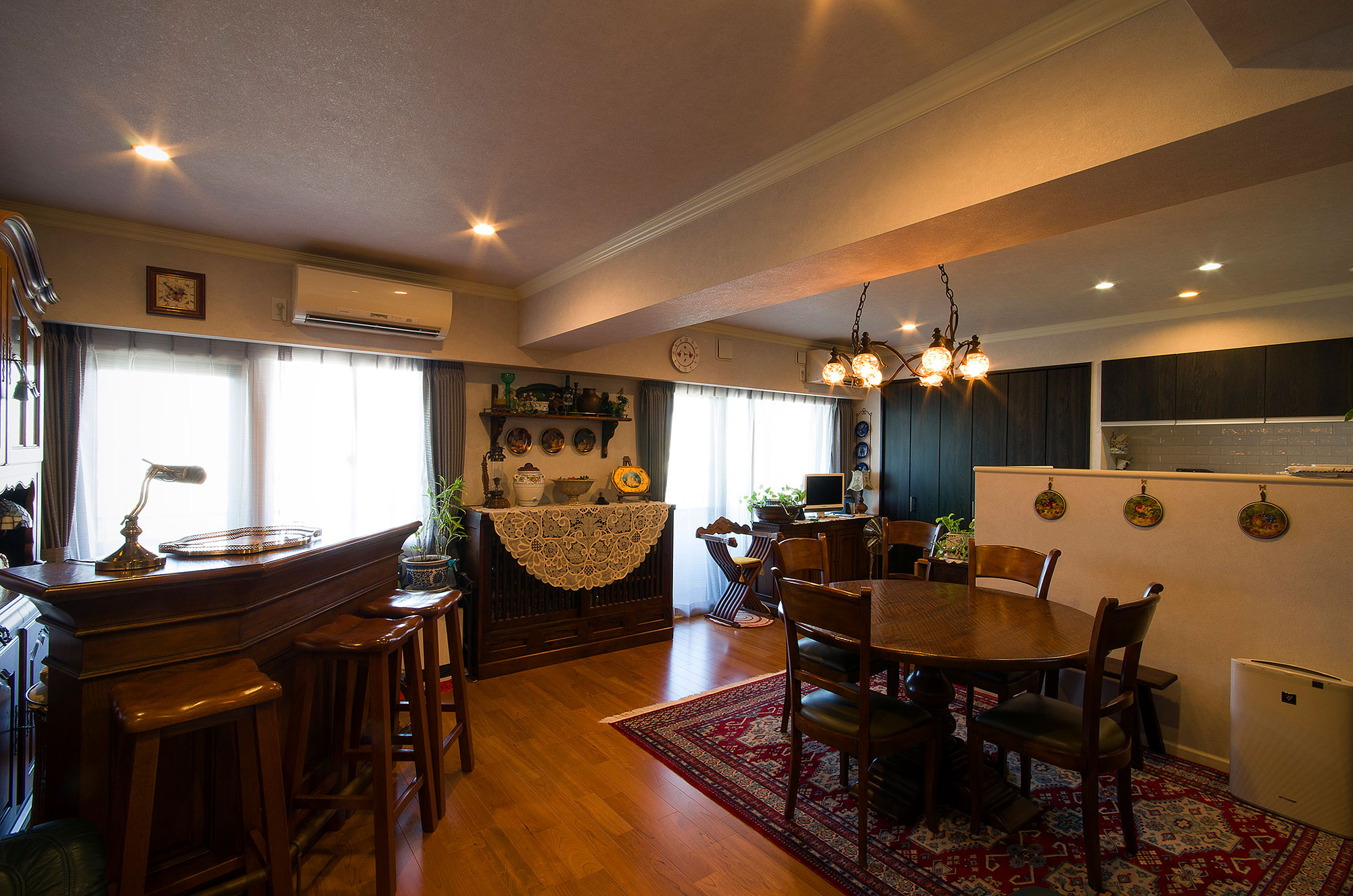 施工写真。重厚なダークブラウンの木製家具で統一された広々とした空間。左手には木製のバーカウンターとスツールが並び、右手には赤い柄物のラグの上に円形のダイニングセットが配置されている。窓際のキャビネットにはレースの敷物やアンティーク雑貨が飾られ、天井のシャンデリアが空間全体を温かく照らしている。奥には白いハーフウォールで仕切られたキッチンが見える。