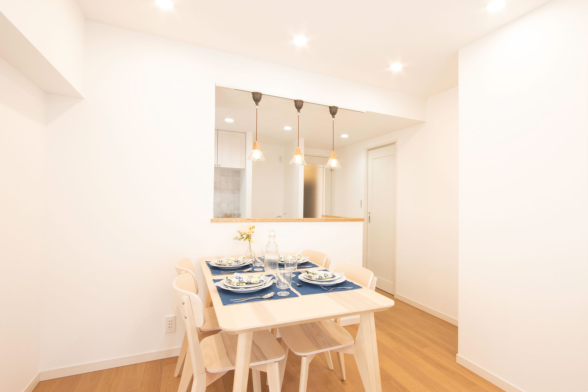 施工写真。白い壁と明るいフローリングの空間に、ナチュラルな木目のダイニングテーブルとチェアが配置されている。テーブルにはブルーのマットと食器がセットされ、奥の対面キッチンからは3つの小ぶりなペンダントライトが下がっている。全体が白を基調とした清潔感のあるコーディネート。