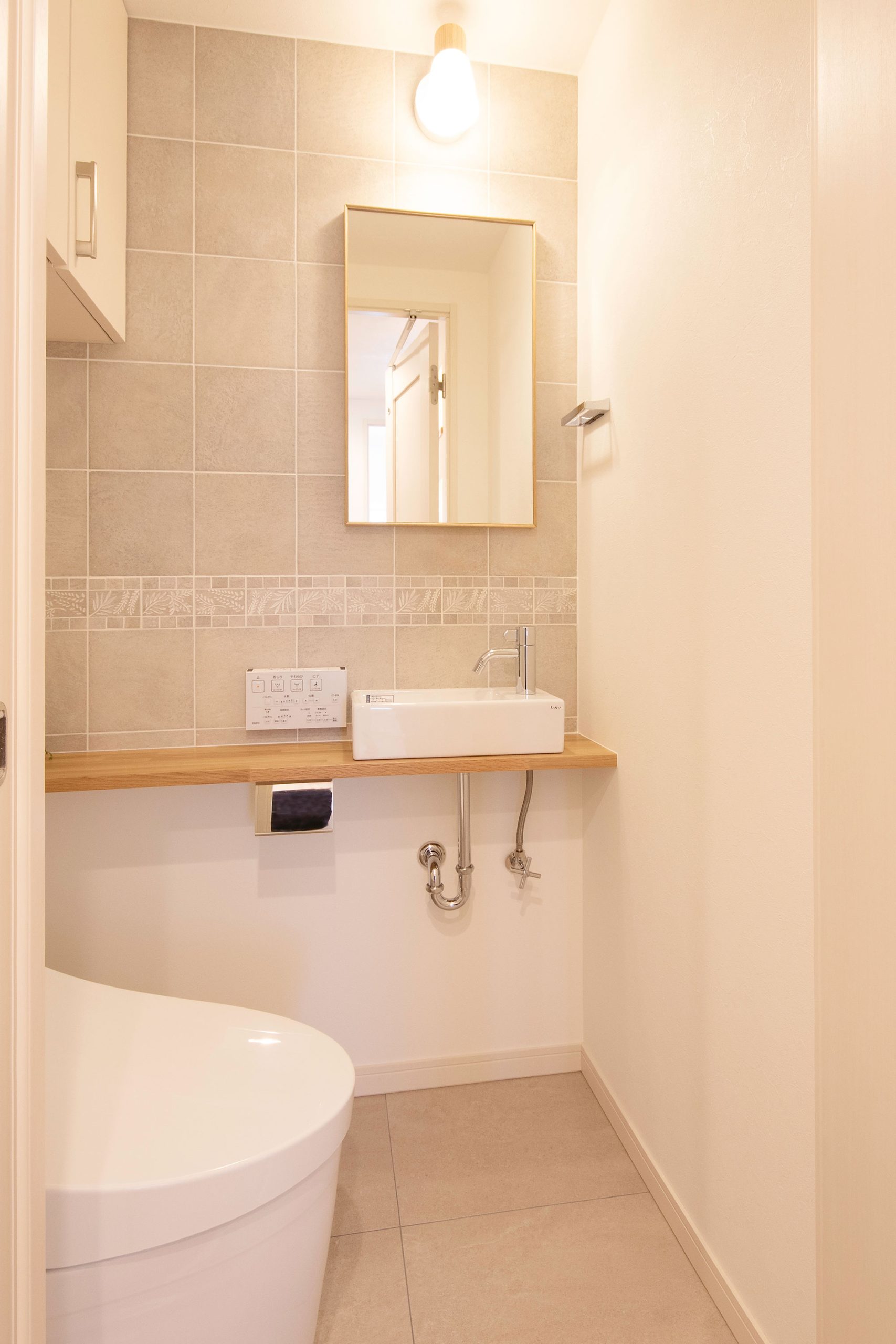 施工写真。白いタンクレストイレの横に、明るい木目のカウンターと四角い手洗いボウルが設置された空間。正面の壁はニュアンスのあるベージュのタイル張りで、繊細な模様のボーダータイルがアクセントになっている。シンプルな縦長ミラーの上には、温かみのある裸電球風のライトが灯っている。