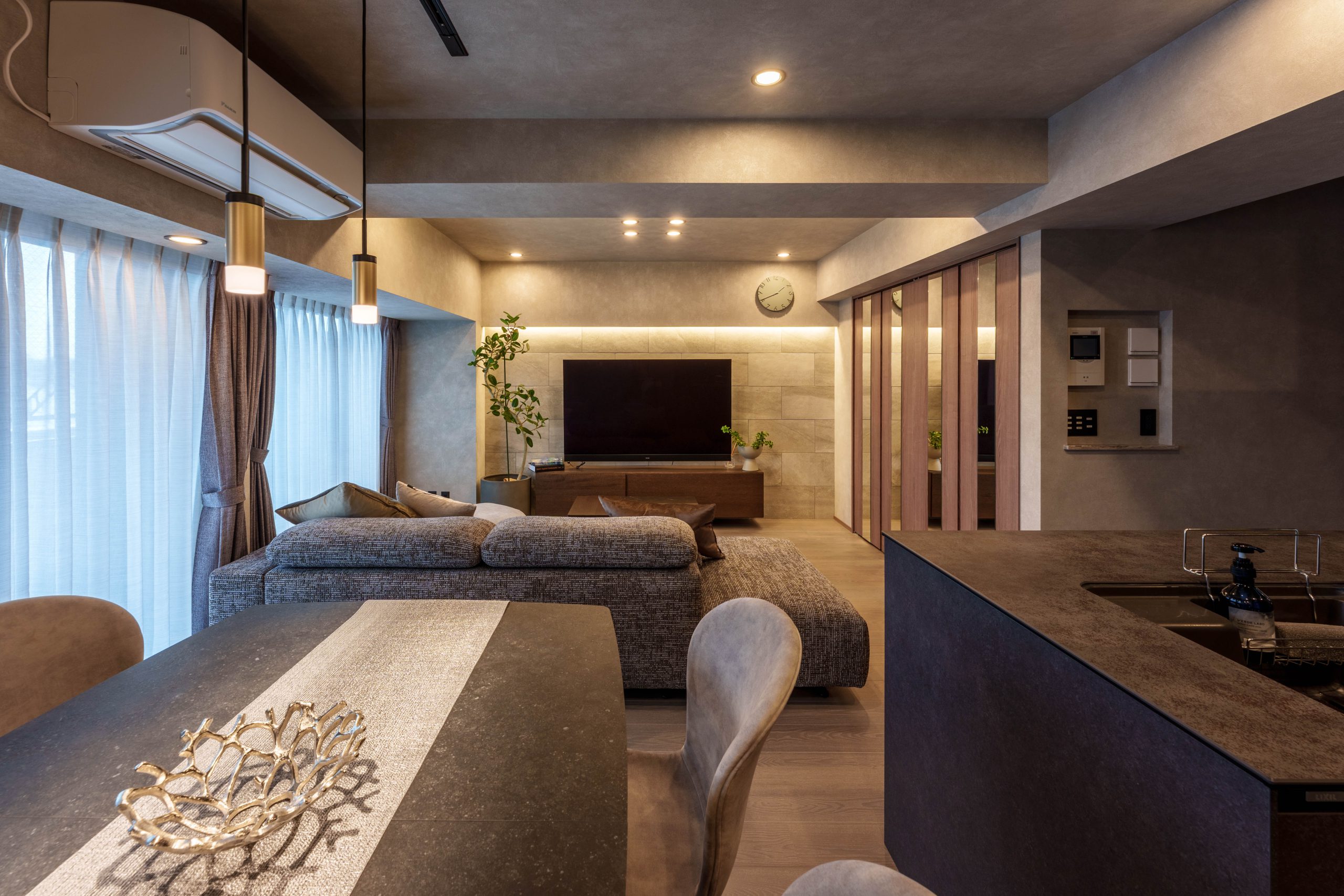 施工写真。ダイニングテーブル、リビングソファ、そしてキッチンを一望するアングル。正面奥のテレビボード背面の壁には、間接照明が石目調タイルの質感を際立たせている。左手の大きな窓からは柔らかな光が入り、天井のダウンライトやゴールドのペンダントライトが、ダークトーンで統一されたインテリアに温かみのある輝きを添えている。